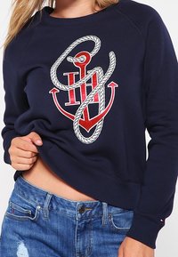 Navy sweatshirt with red anchor and white rope design, worn by person lifting hem, paired with blue distressed jeans.