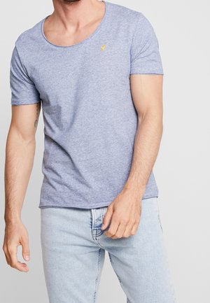 Homme portant un t-shirt bleu clair à col rond avec un petit logo jaune sur la poitrine et un jean bleu clair, debout devant un fond uni.
