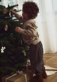 Dítě v béžovém pleteném svetru s motivem stromů zdobí zelený vánoční stromeček dřevěnými a papírovými ozdobami. Hnědé kordové kalhoty a boty.