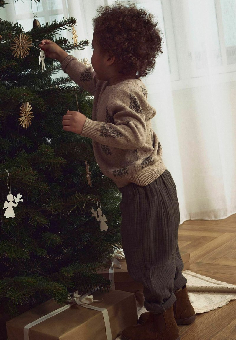 Dítě v béžovém pleteném svetru s motivem stromů zdobí zelený vánoční stromeček dřevěnými a papírovými ozdobami. Hnědé kordové kalhoty a boty.