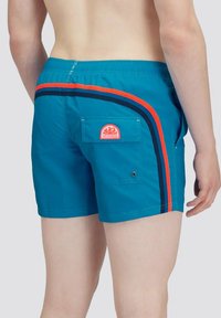 Pantaloni da bagno turchesi con un accento a strisce rosse e navy. Presentano una tasca posteriore con chiusura a scatto e un patch con il logo. Tessuto leggero, vestibilità atletica.