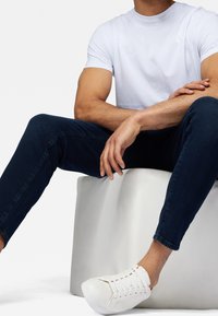 Vit bomullströja, mörkblå jeans och vita sneakers med en strukturerad yta; person sitter på en vit kub.