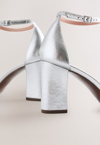 Paire de sandales argentées à talons blocs avec brides à la cheville et petites boucles métalliques sur fond clair et neutre.