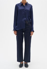 Camicia lunga in satin blu marina con chiusura a bottone, abbinata a jeans scuri a gamba larga e scarpe nere slip-on. Design semplice e moderno.