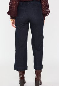 Pantaloni in denim scuro a vita alta, con gambe larghe e orlo arrotolato, dotati di due tasche posteriori. Indossati con stivali marroni con tacco.