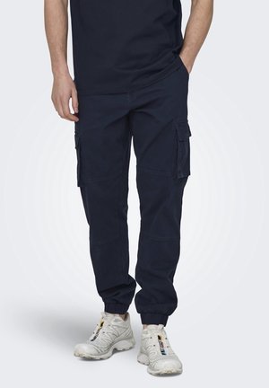 Pantaloni cargo della Marina con polsini elasticizzati; presentano molteplici tasche laterali e una vestibilità comoda. Indossati con scarpe da ginnastica di colore chiaro.