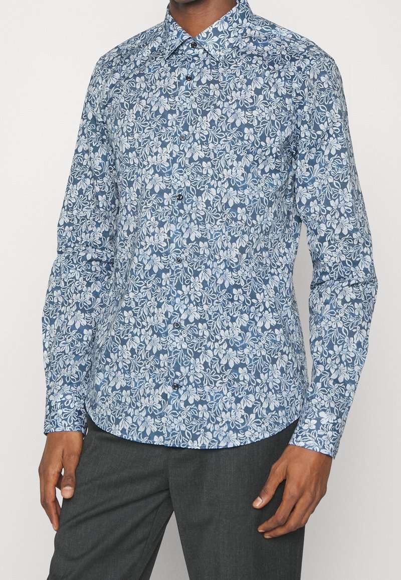 Homme portant une chemise bleue à manches longues avec un motif floral blanc et un pantalon gris foncé, debout devant un fond blanc.