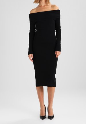 Mujer con vestido midi negro ajustado con escote barco, mangas largas y tacones negros puntiagudos sobre un fondo liso.