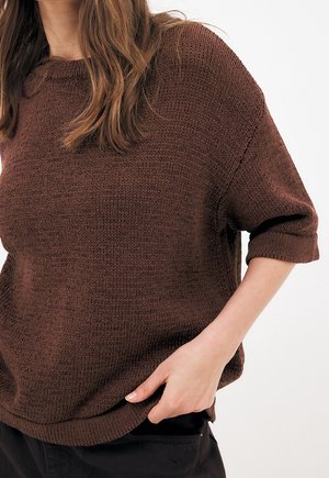 Vrouw draagt een losse bruine gebreide trui met opgerolde mouwen, hand in de zak van een zwarte broek, weergegeven van nek tot taille.
