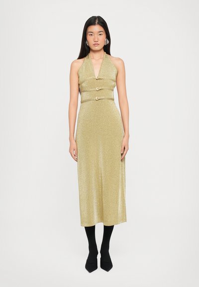 Goldenes Halsbandkleid mit tailliertem Oberteil, drei dekorativen Riemen und Metallverschlüssen, knielang, strukturiert, getragen mit schwarzen Strumpfhosen und hohen Schuhen.