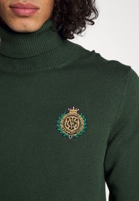 Dunkelgrüner Rollkragenpullover aus Strickstoff mit einem runden, goldenen und blauen gestickten Logo mit Krone und Blättern auf der Brust.