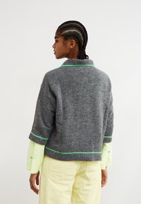 Maglione grigio lavorato a maglia con collo corto, caratterizzato da dettagli verdi lungo i bordi. Il modello indossa una camicia giallo chiaro e strutturata sotto.