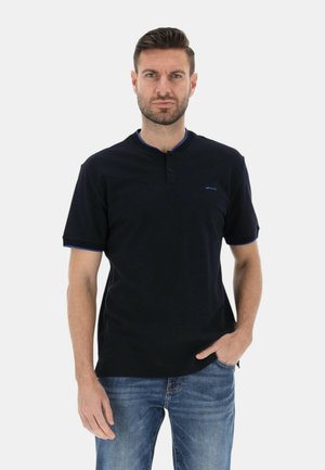 Uomo che indossa una maglia nera a maniche corte con rifiniture blu e jeans blu, in piedi con una mano in tasca su sfondo bianco.