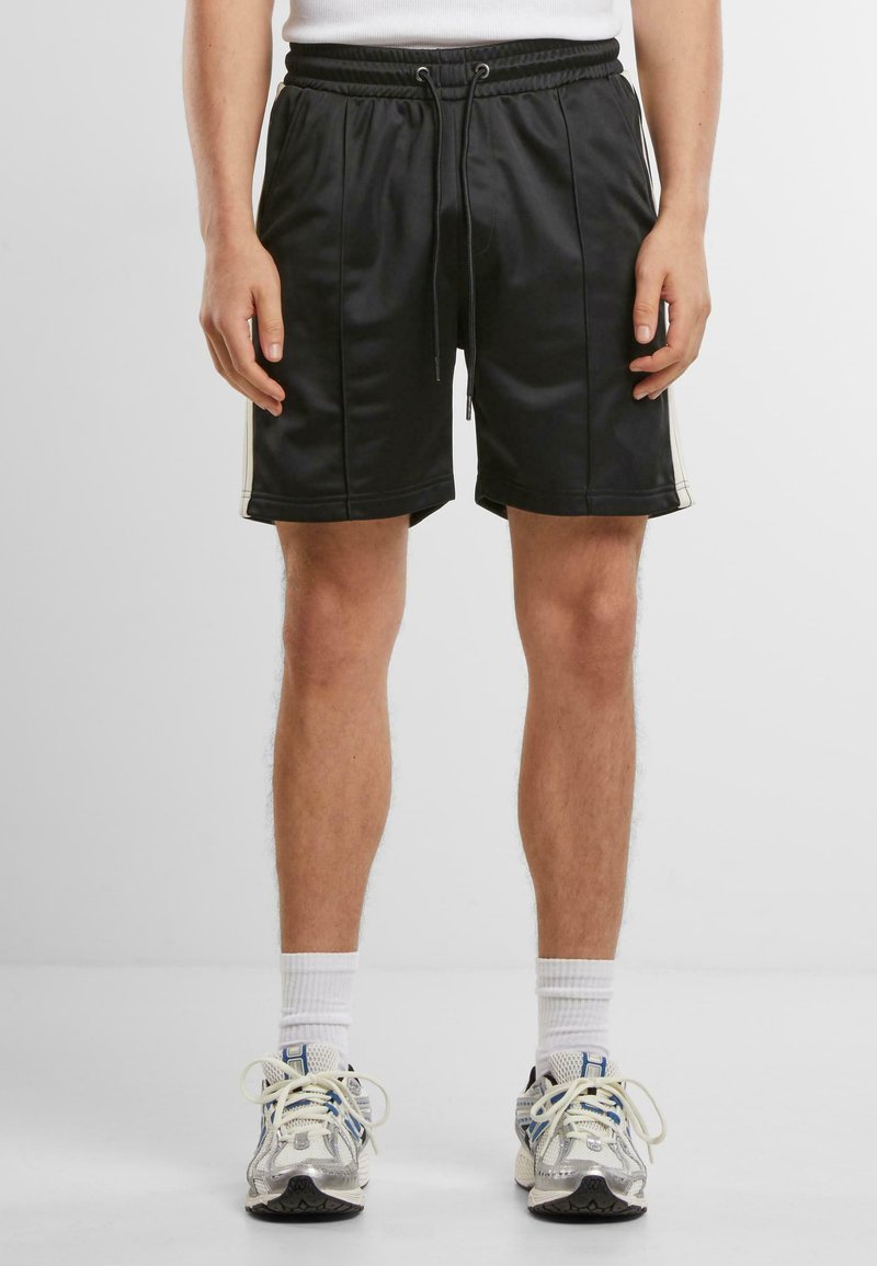 Shorts de sport noirs avec une taille à cordon, présentant des bandes latérales, une texture lisse et un design jusqu'aux genoux. Portés avec des chaussettes blanches et des baskets.