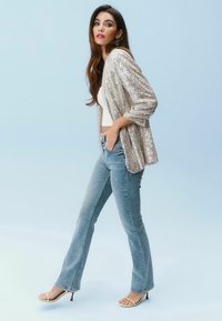 Silberne Pailletten-Cardigan, weißes Crop-Top, hellblaue Schlagjeans und Riemchensandalen. Das Outfit hat ein geschichtetes, texturiertes Aussehen.
