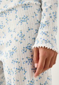 Κοντινό πλάνο χεριού που ακουμπά σε λευκό ύφασμα με μικρό μπλε λουλουδένιο σχέδιο και λεπτομέρεια τελείωμα με κυματιστό άκρο στο μανίκι.