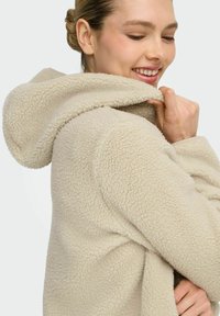 Beige fleecejakke med hette, teksturert stoff og ledig passform. Materialet har et mykt, plysjaktig utseende med synlige sømmer.