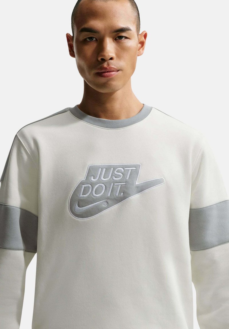 Primer plano de una persona que lleva un sudadera blanca y gris con el logo de Nike y el texto "JUST DO IT." en el pecho.