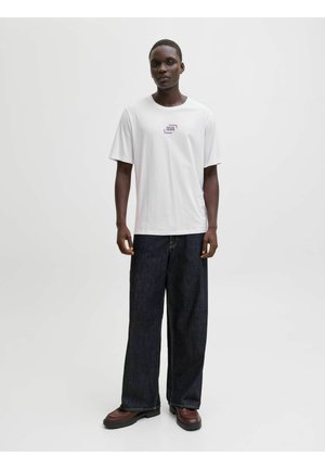 Person steht, trägt ein weißes Jack & Jones T-Shirt, weite dunkle Jeans und braune Schuhe vor einem schlichten hellen Hintergrund.