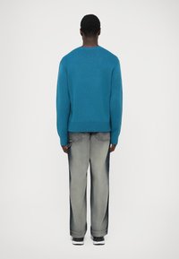 Ein Mann steht mit dem Rücken zur Kamera, trägt einen türkisfarbenen Strickpullover, ausgewaschene, lockere Jeans mit dunklen Seitenpanelen und schwarz-weiße Sneakers vor einem weißen Hintergrund.