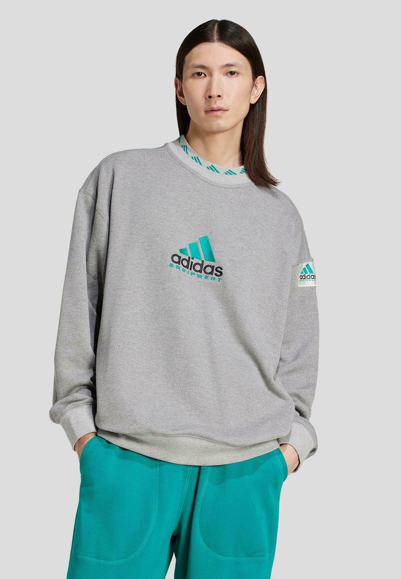 Grå sweatshirt med teal-accenter och broderad logotyp, med en lös passform och ribbat halsband. Parat med tealbyxor.