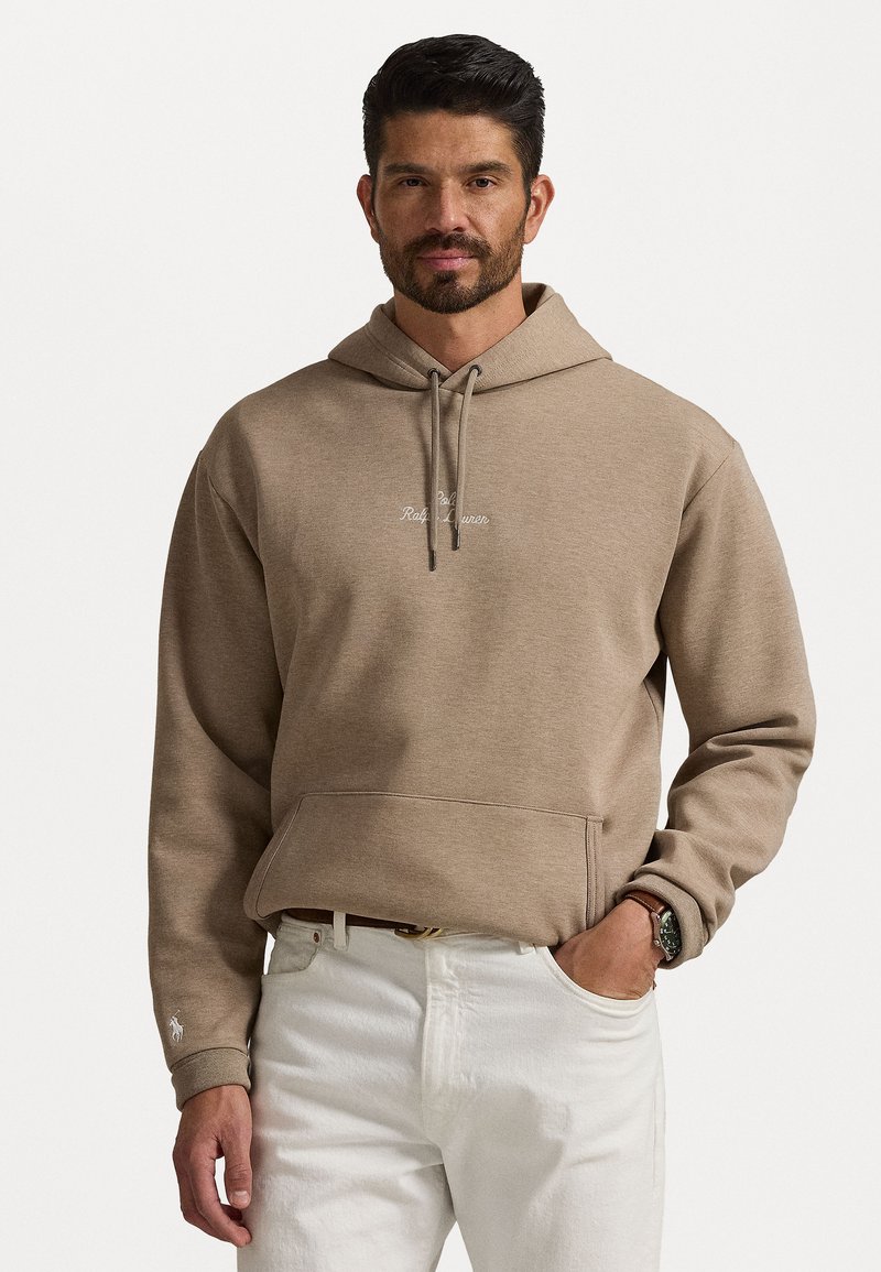 Felpa in cotone color cammello con tasca frontale, dotata di cordini e un piccolo logo ricamato sul petto. Abbinate a pantaloni bianchi.