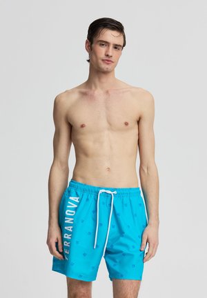 Giovane uomo in piedi a torso nudo che indossa pantaloncini da bagno azzurri vivaci con coulisse bianca e testo "TERRANOVA" sulla gamba sinistra.