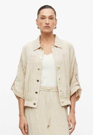 Femme aux cheveux plaqués en arrière portant une veste beige boutonnée, un haut blanc et une jupe beige assortie à cordon, sur fond blanc uni.