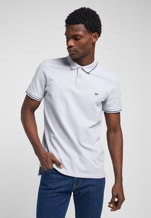 Mężczyzna w jasnoszarej koszulce polo z granatowym obszyciem i logo "Lee", w niebieskich dżinsach, stojący na jednolitym szarym tle.