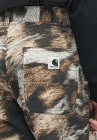 Πίσω τσέπη καφέ, μαύρων και λευκών παντελονιών tie-dye με λογ�ότυπο Carhartt, φορεμένα από άτομο με μαύρη μακρυμάνικη μπλούζα.