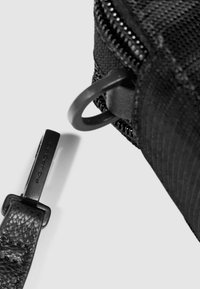 Primo piano di una borsa in tessuto nero testurizzato con una cerniera e una fibbia metallica, insieme al dettaglio dell'hardware di una tracolla in pelle.