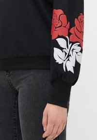Svart hettegenser med rød rose og hvit bladdesign, kombinert med mørk grå jeans og en avslappet hånd ved siden.