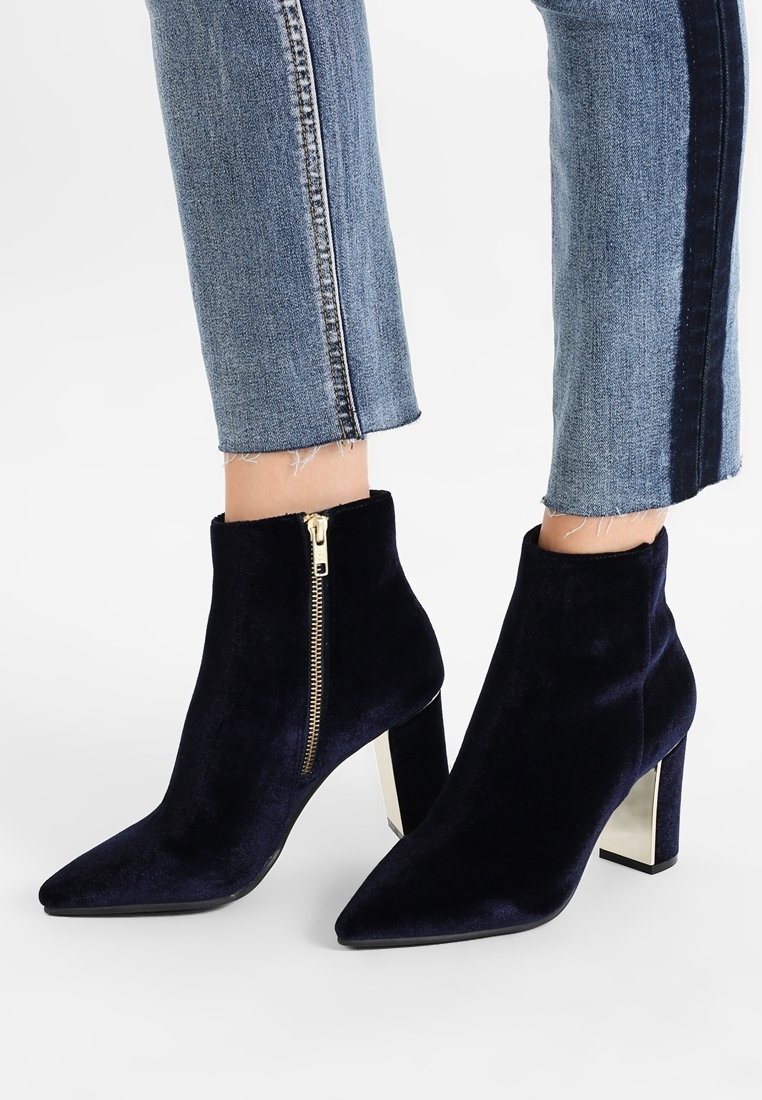 Botines de terciopelo azul marino hasta el tobillo con punta afilada, detalle de cremallera dorada y tacón metálico grueso. Textura suave con una silueta elegante.