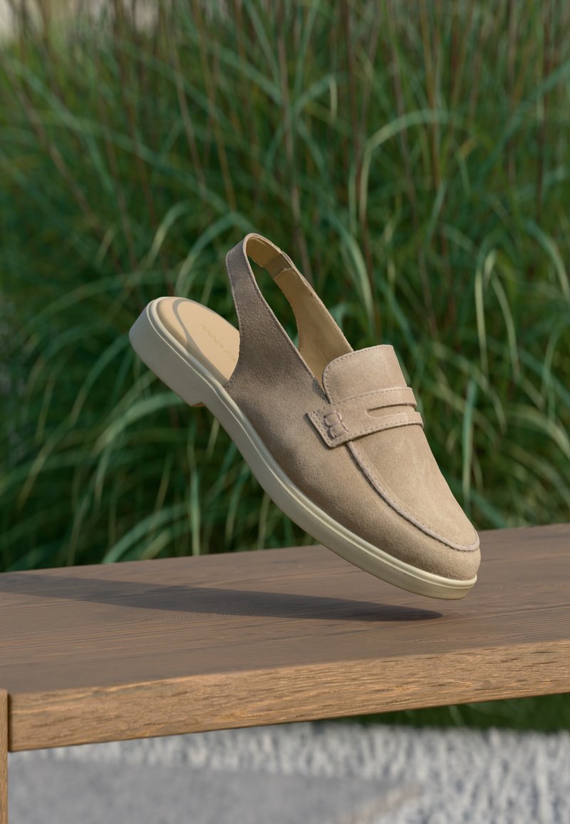 Mocassin slingback beige en daim avec semelle blanche reposant sur un banc en bois sur un fond d’herbes hautes et vertes.