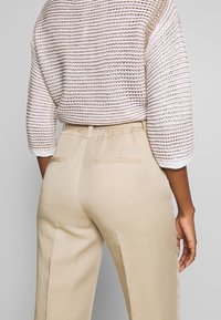 Beige linnen broeken met zijzakken en riemlussen, gecombineerd met een losse, gebreide crèmekleurige trui met horizontale strepen.