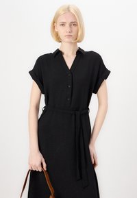 Rochie neagră dintr-un material cu textură, cu un decolteu tip cămașă, mâneci scurte rulate, talie legată și un buzunar în față. Este însoțită de o geantă maro.