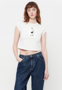 Witte cropped t-shirt met een zwart-wit grafisch ontwerp. Gecombineerd met losse donkerblauwe spijkerbroek met een lichte glans en zichtbare stiksels.