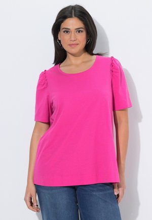 Kvinde iført en skrigende pink kortærmet top med rynkede skulderdetaljer og blå jeans, stående op ad en hvid væg.