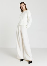 Weißer Strickpullover mit strukturierten Mustern, geripptem Saum und langen Ärmeln, kombiniert mit hochgeschnittenen weißen weitem Hosen und schwarzen spitzen Absätzen.