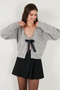 Kobieta poprawiająca szary sweter z dzianiny z perełkami i czarną kokardą, ubrana w czarną plisowaną spódnicę i kolczyki-koła, uśmiechająca się na białym tle.