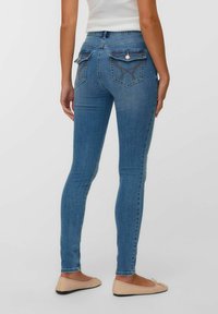 Sieviete, ģērbusies vidēji zilās šaurajās biksēs ar aizsargpogu aizmugurējām kabatām un smilšu krāsas balerīnām, stāv pretī vienkāršai gaišai fona.