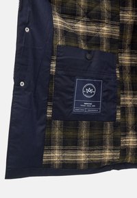 Navy takki sisäosa, jossa on vihreä- ja beigevärinen ruudullinen kuvio. Mukana on tasku, jossa on tuotelogo, joka sisältää bränditietoja.