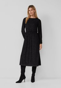 Kobieta w czarnej, teksturowanej midi sukience z długim rękawem i dopasowanym w pasie, w czarnych, wysokich na kolano, obcasach, stojąca na jednolitym tle.