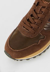 Sneaker en daim marron et cuir avec des lacets assortis et une semelle intermédiaire beige sur un fond blanc.