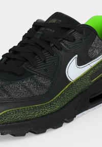 Μαύρο αθλητικό παπούτσι Nike Air Max με λευκό swoosh, υφασμάτινες γκρι και πράσινες λεπτομέρειες, μαύρα κορδόνια και νέον πράσινη εσωτερική επένδυση.