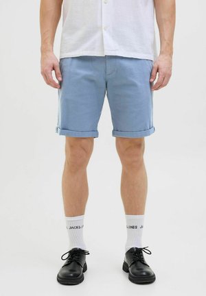 Pantaloni corti in cotone azzurro chiaro con orli arrotolati, abbinati a scarpe nere e calzini bianchi con testo. La maglietta è bianca e leggermente comoda.