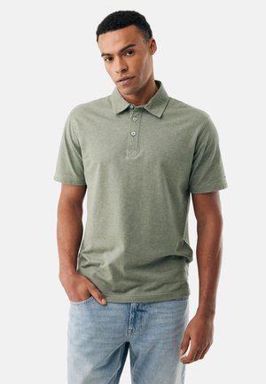 Mann trägt ein grünes kurzärmeliges Poloshirt und hellblaue Jeans, steht mit einer Hand in der Tasche vor einem schlichten weißen Hintergrund.