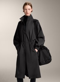 Kvinna med kort hår iförd en lång svart zip-up kappa med hög krage, händerna i fickorna, bärande en stor svart axelväska.