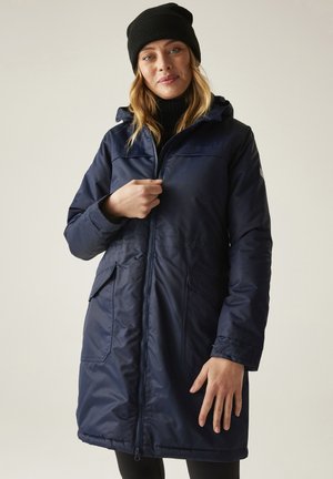 Donna che indossa un cappotto invernale con cappuccio blu scuro e un berretto lavorato a maglia nero, che chiude la cerniera frontale contro uno sfondo beige uniforme.