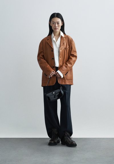 Brown leather jacket with black buttons, white shirt, black wide-leg trousers, and black handbag. Model wearing black lace-up boots.